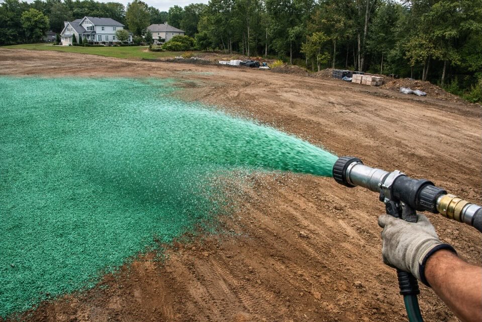 Aerial view of a green hydroseeding slurry being sprayed from a professional hose nozzle over a large graded residential lot in New Jersey, lush green mulch coating bare brown soil, overcast natural lighting.
